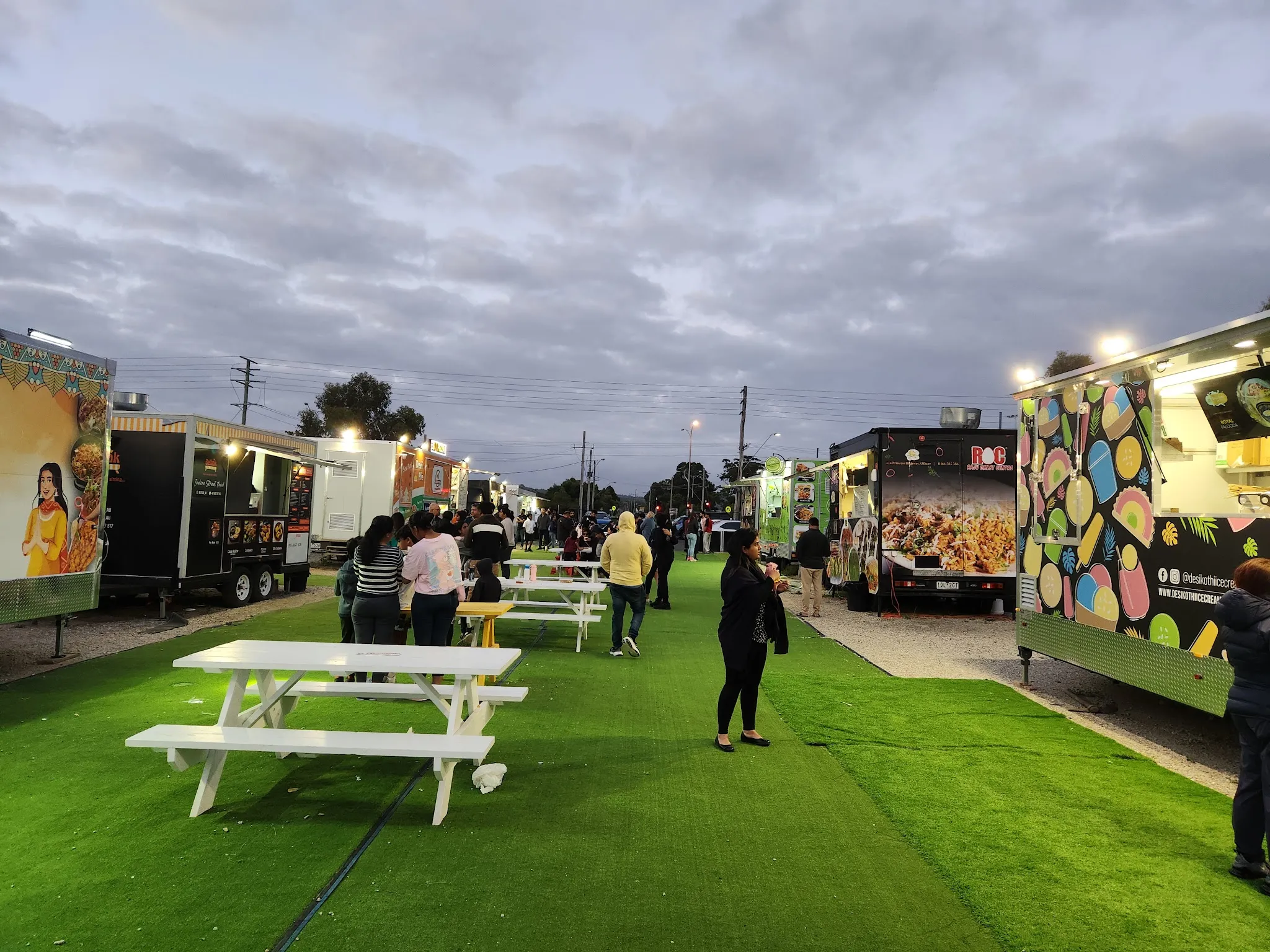 Wide dusk view down the central lane of Officer Food Truck Park, trucks strung with lights and picnic tables filling with families.