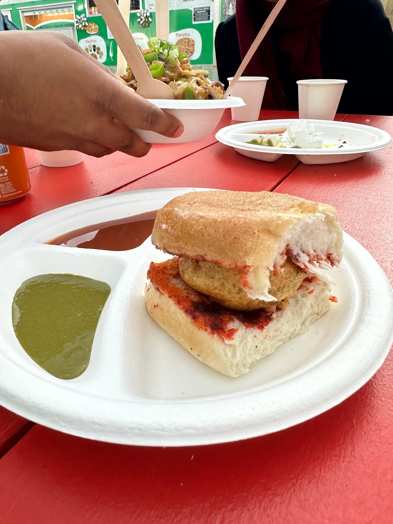 A vada pav sandwich held over a tray of chutneys with other dishes visible on the table.