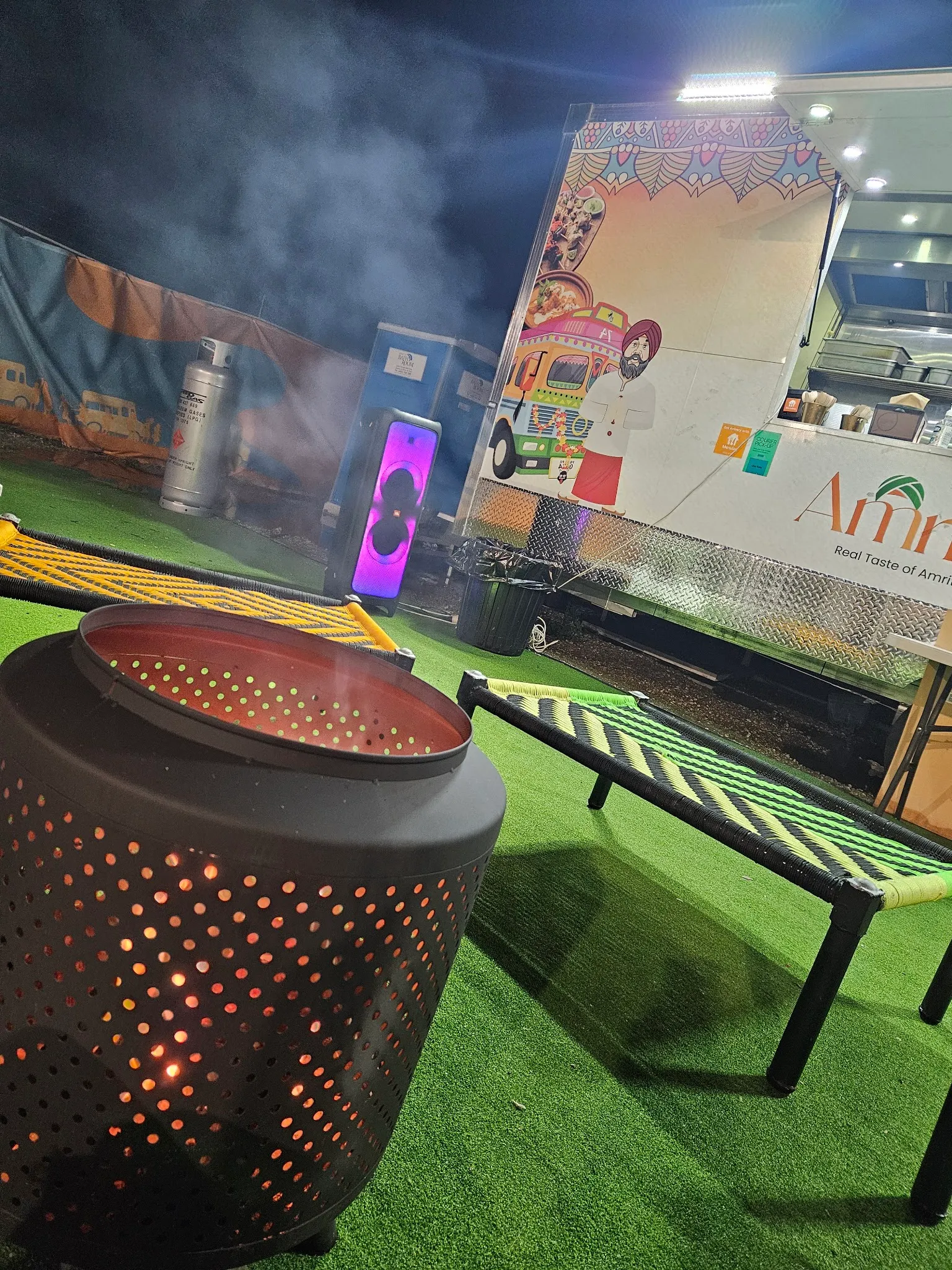 The Amritsari food truck at night with illustrated signage and light haze drifting from a nearby fire pit.