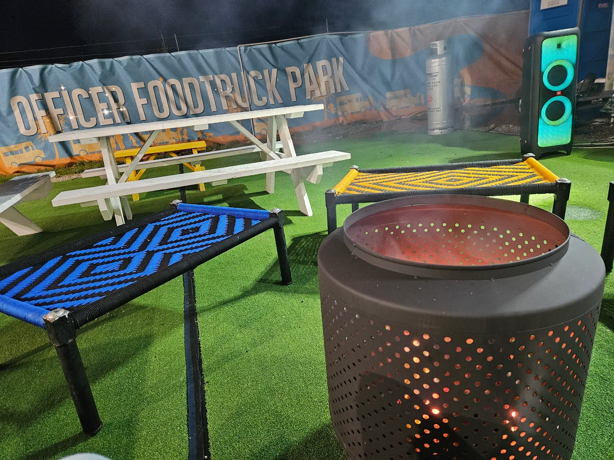 A nighttime lounge corner of the park with a glowing fire pit and woven benches beneath a lit-up truck banner.